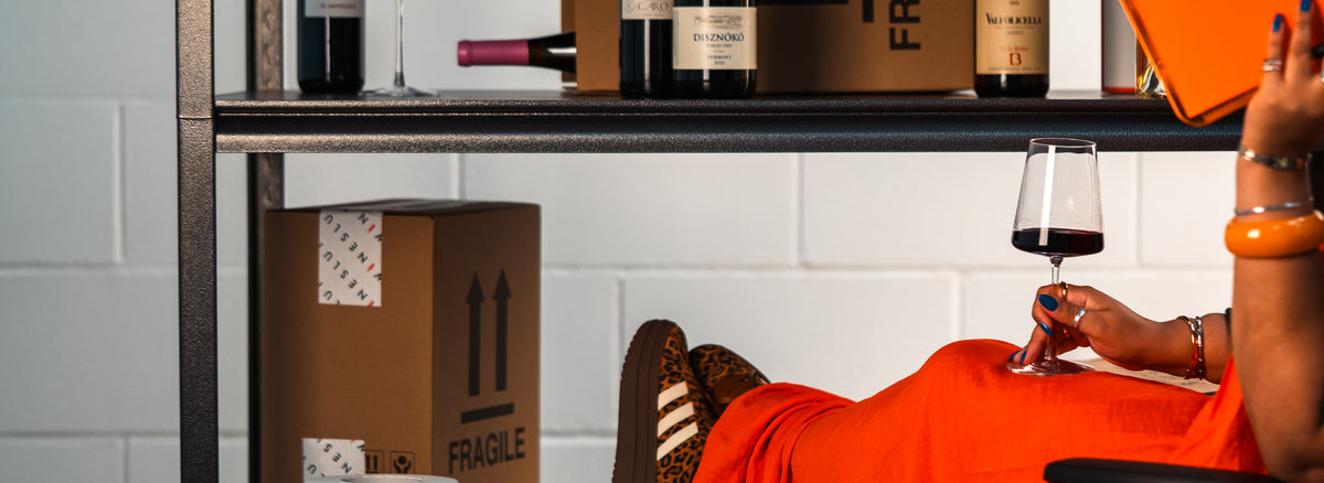 A person in an orange dress reclines on a shelf while holding a glass of red wine, with cardboard shipping boxes and wine bottles stacked on a metal shelving unit in the background.