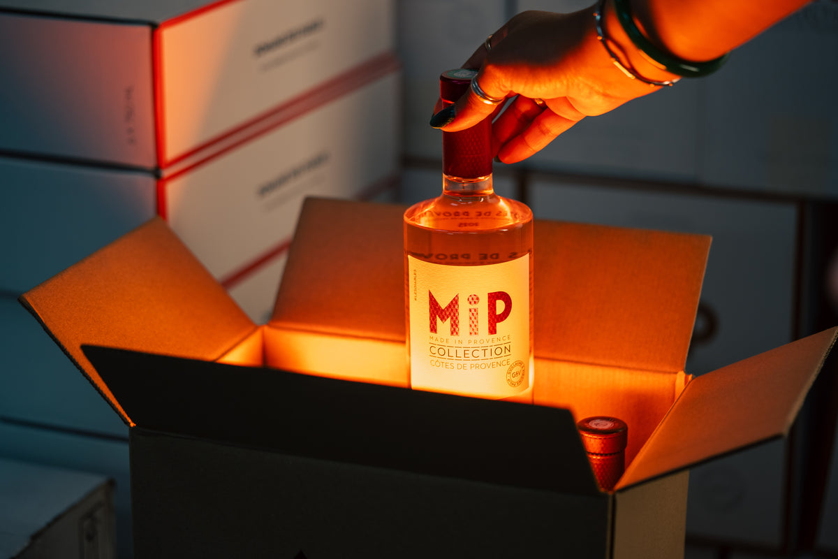 A hand holds a bottle of MiP Collection Rose from a brown shipping box with orange lighting, with stacked boxes in the background.