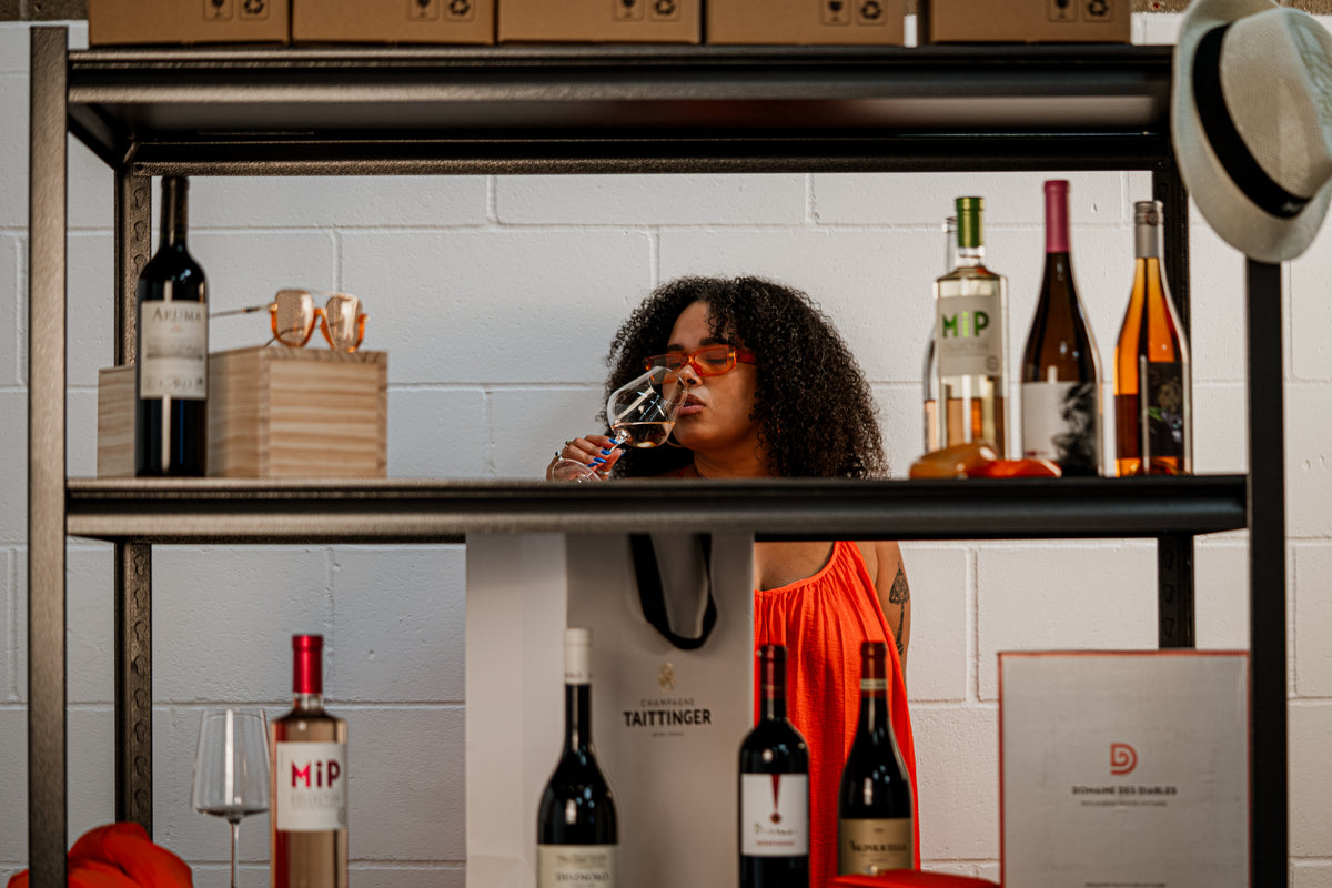 A woman with curly hair wearing an orange top and sunglasses peers over a wine rack, holding a glass of red wine. The rack is stocked with various wine bottles, glasses, and boxes, set against a white brick wall.