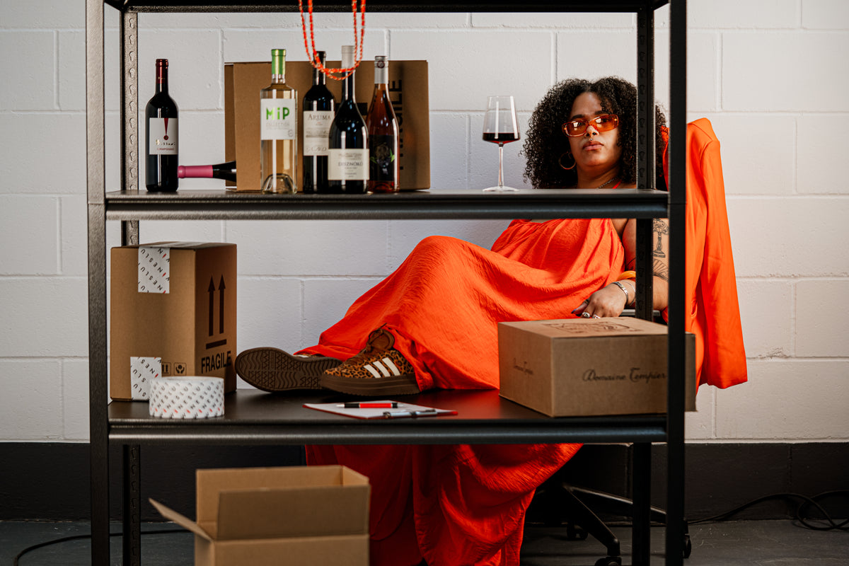 A person reclining on a black metal shelving unit in a room with a white brick wall, wearing a bright orange dress and orange-tinted sunglasses. The shelves are styled with wine bottles, a glass of red wine, cardboard boxes, and a brown Adidas sneaker.